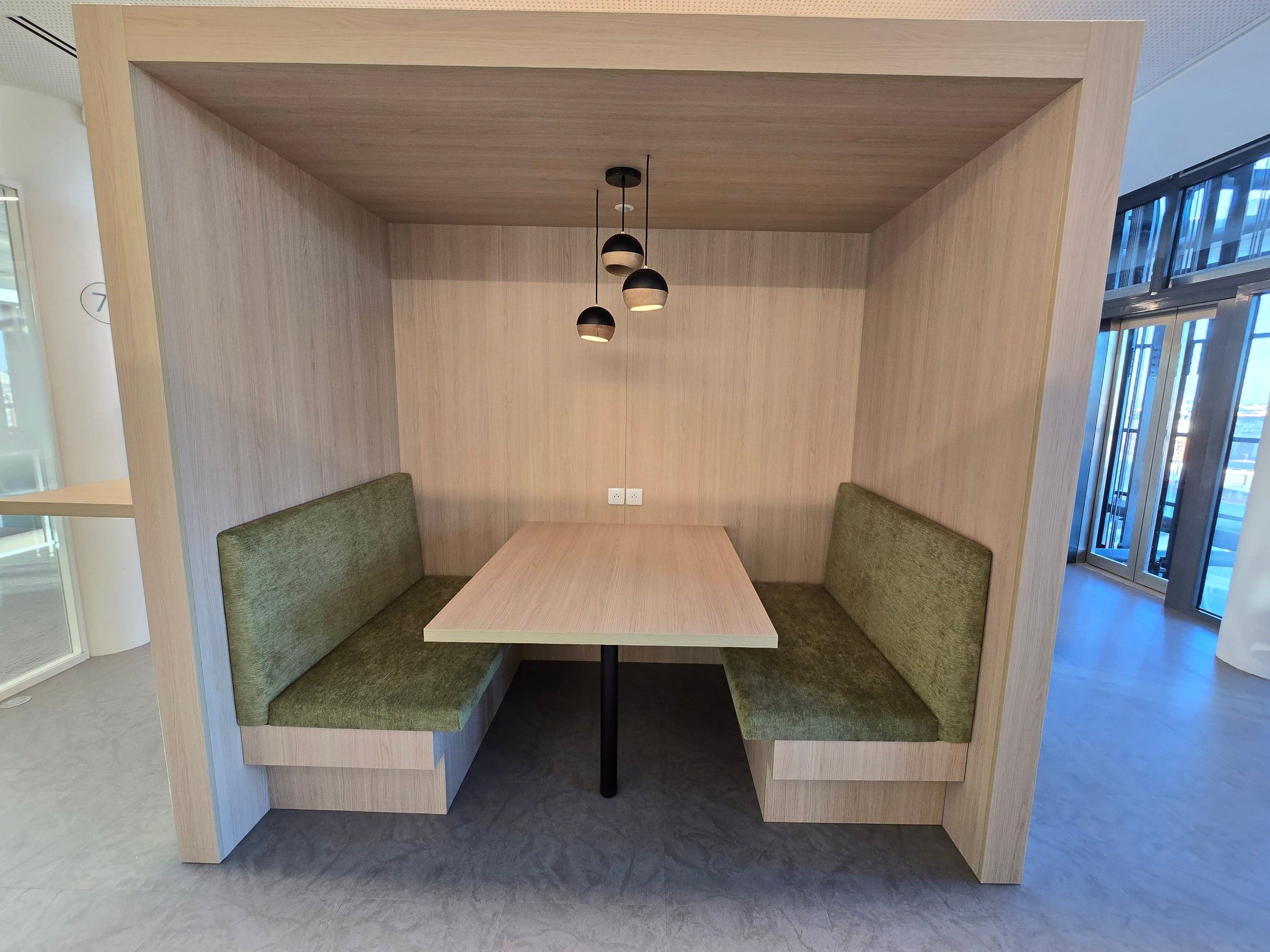 Alcôve en décor bois clair avec deux banquettes vertes face à face, une table et trois suspensions