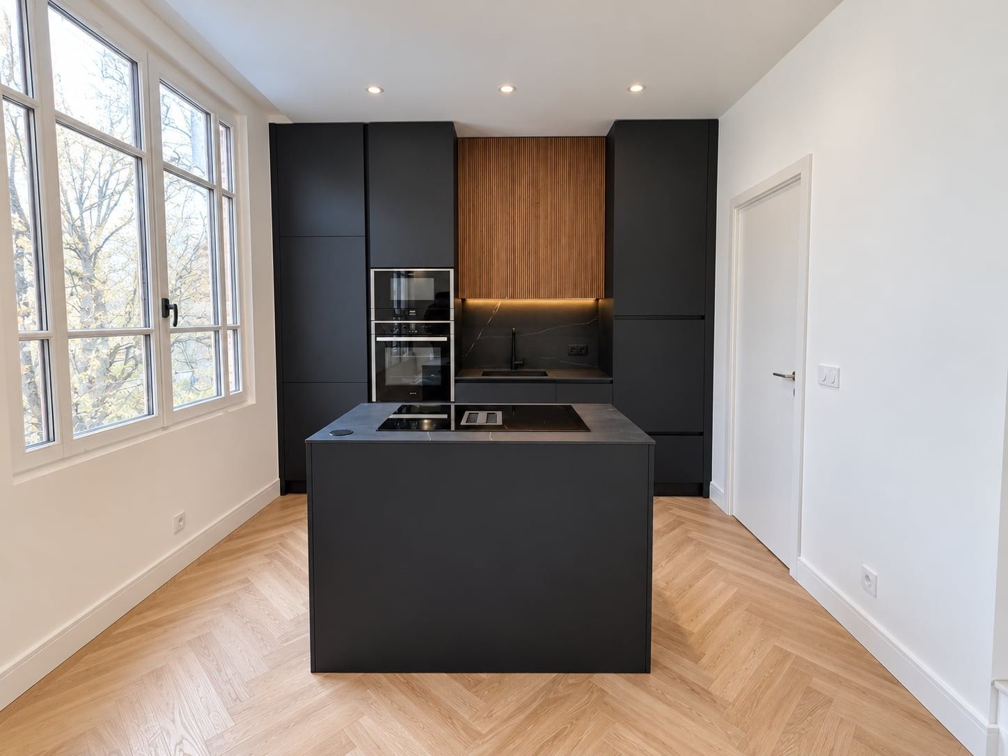 Cuisine noir mat vue de face : îlot central, colonnes, caissons en bois rainuré et grandes fenêtres à gauche
