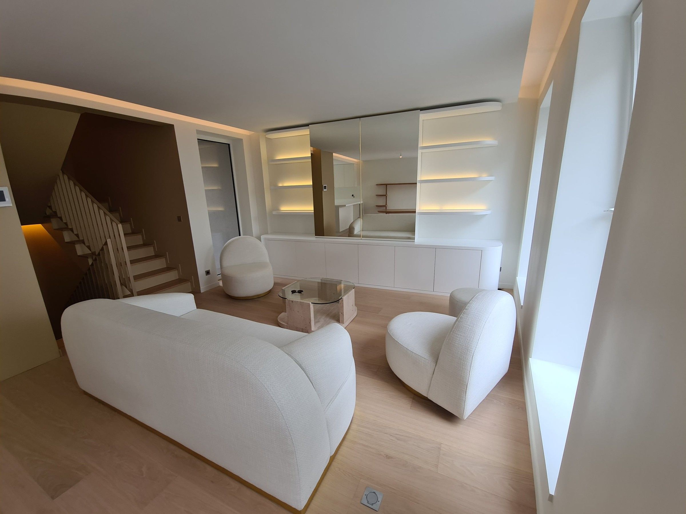 Séjour blanc avec canapé et fauteuils arrondis, meuble bas cintré et étagères lumineuses autour d'un miroir