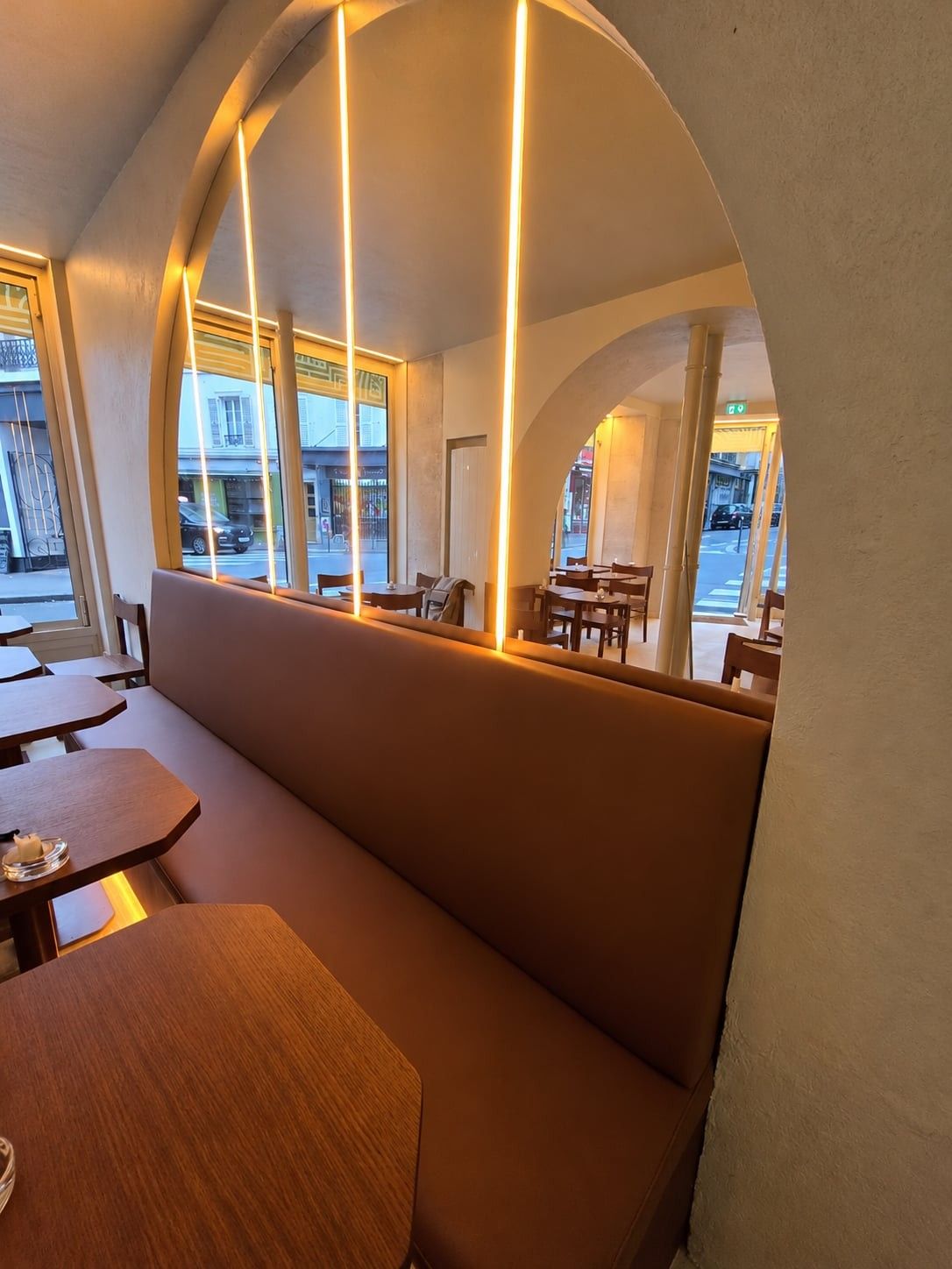 Banquette camel à haut dossier sous une arche éclairée de baguettes LED, face aux tables en bois