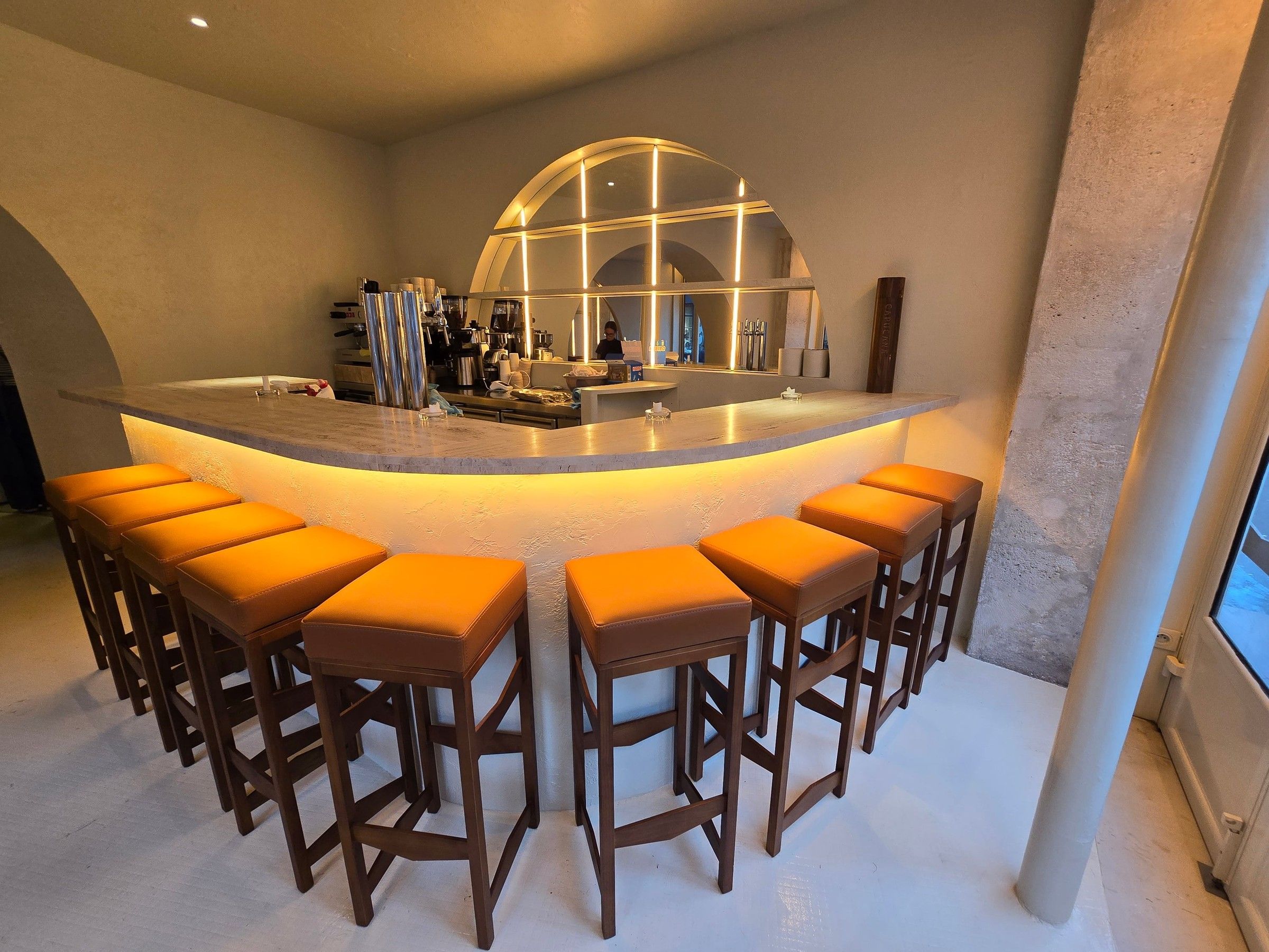 Bar cintré éclairé sous son plateau en pierre, bordé de tabourets en bois à assise camel, devant une arche lumineuse