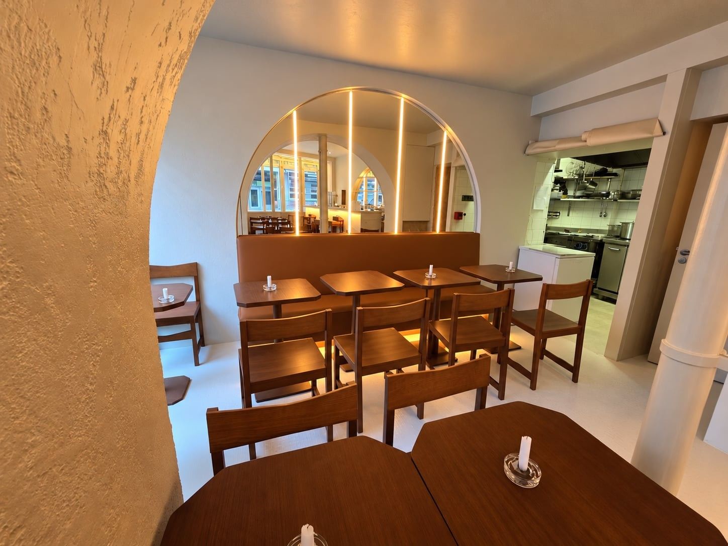 Salle avec banquette murale camel, tables à pans coupés et chaises en bois, sous une arche ouverte vers le bar